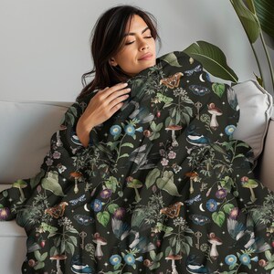 May include: A dark green throw blanket featuring a whimsical pattern of flowers, birds, mushrooms, and butterflies. The blanket is soft and cosy, ideal for snuggling up on the sofa.