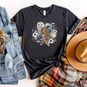 May include: A dark gray t-shirt with a white barn owl, flowers, and a pocket watch design. The owl is surrounded by white flowers and green leaves. The pocket watch has Roman numerals and a gold chain.