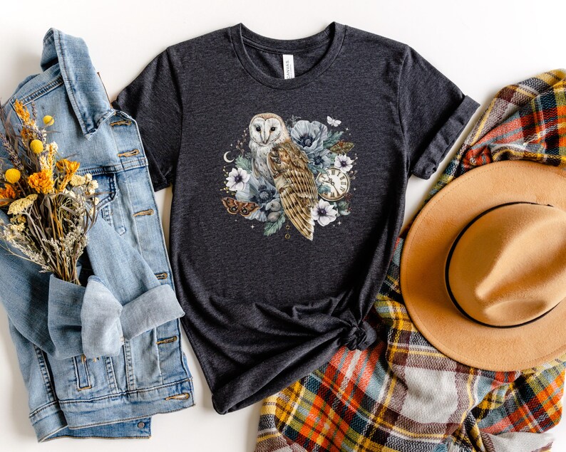 May include: A dark gray t-shirt with a white barn owl, flowers, and a pocket watch design. The owl is surrounded by white flowers and green leaves. The pocket watch has Roman numerals and a gold chain.