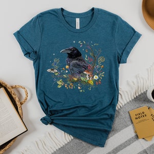 May include: Teal blue t-shirt with a black raven illustration surrounded by colorful flowers and mushrooms.