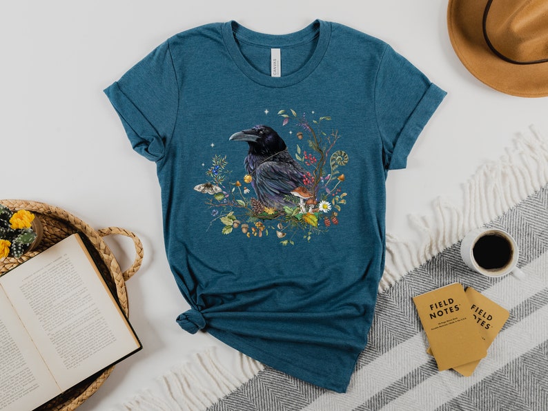 May include: Teal blue t-shirt with a black raven illustration surrounded by colorful flowers and mushrooms.