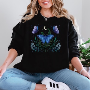 Puede incluir: Sudadera negra con una mariposa azul, luna y diseño floral. El diseño presenta una gran mariposa azul con detalles florales verdes y blancos.