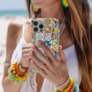 Peut inclure: Coque de téléphone colorée avec un motif patchwork composé de motifs floraux et géométriques bleus, jaunes, rouges et blancs. La coque est tenue devant une personne, avec une plage en arrière-plan.