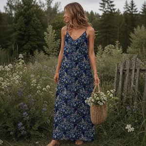 May include: A long, flowing dress with a blue floral pattern. The dress has a V-neck and spaghetti straps. A woven basket filled with white flowers is held in the hand. The background features a field of wildflowers and trees.