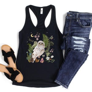 May include: Black racerback vest top with a nature-themed graphic of a white owl, flowers, and foliage. Shown with dark wash denim jeans and black bow sandals. A casual, bohemian style.