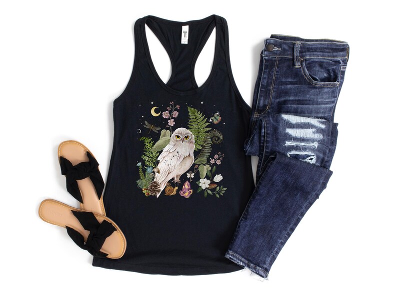 May include: Black racerback vest top with a nature-themed graphic of a white owl, flowers, and foliage. Shown with dark wash denim jeans and black bow sandals. A casual, bohemian style.