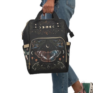 Può includere: Zaino nero con un design a farfalla e luna. Lo zaino ha una cerniera dorata e una tracolla dorata. Il testo sullo zaino recita "perfect phase to shine".