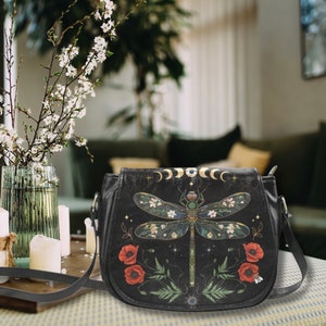 May include: A black leather crossbody bag with a dragonfly design. The dragonfly is surrounded by flowers and leaves. The bag has a gold chain strap and a zipper closure.