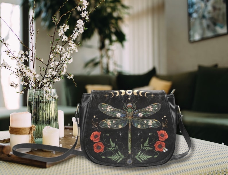 May include: A black leather crossbody bag with a dragonfly design. The dragonfly is surrounded by flowers and leaves. The bag has a gold chain strap and a zipper closure.