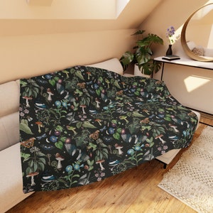 May include: A dark blanket with a botanical pattern featuring mushrooms, butterflies, birds, and keys. The design includes green leaves, blue flowers, and other natural elements. The blanket is draped over a beige sofa.