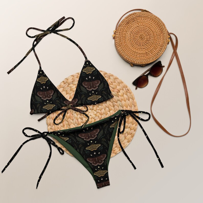 May include: A black and green bikini with a moth print. The bikini top is a triangle style and the bottom is a tie-side style. The bikini is on a straw mat with a straw bag and sunglasses.