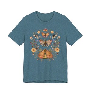 May include: A heathered blue short-sleeve t-shirt featuring a central graphic print of three moths arranged vertically, surrounded by a circular arrangement of orange flowers, small green leaves, and tiny sparkling stars in a symmetrical design.