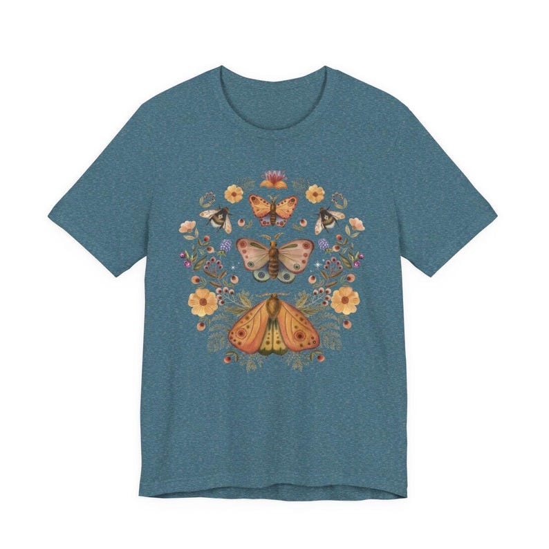 May include: A heathered blue short-sleeve t-shirt featuring a central graphic print of three moths arranged vertically, surrounded by a circular arrangement of orange flowers, small green leaves, and tiny sparkling stars in a symmetrical design.