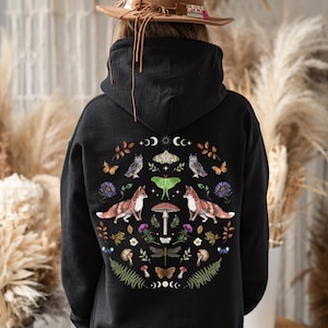 Pode incluir: Camisola de capuz preta com um design circular que apresenta raposas, corujas, borboletas, cogumelos e outras criaturas da floresta. O design é rodeado por uma lua crescente e estrelas.