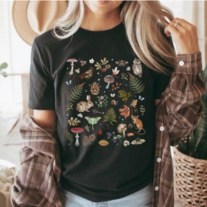 May include: Black t-shirt with a colorful print of various woodland creatures, plants, and mushrooms. The print includes a rabbit, a moth, a bird, a snail, a mushroom, a pine cone, and a mouse.