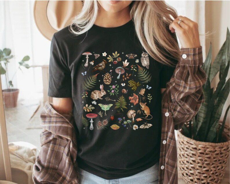 May include: Black t-shirt with a colorful print of various woodland creatures, plants, and mushrooms. The print includes a rabbit, a moth, a bird, a snail, a mushroom, a pine cone, and a mouse.