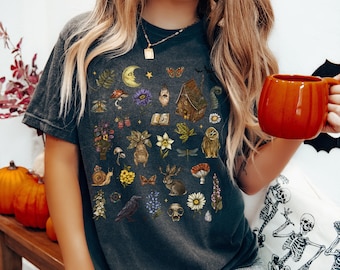May include: Dark gray t-shirt featuring a whimsical illustration of various botanical and mystical elements. The design includes a crescent moon, flowers, a house, an owl, and a rabbit. The shirt is a comfortable, casual style.