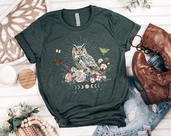 May include: Dark green t-shirt featuring a watercolor owl design with floral and moon phase elements. The owl is the focal point, surrounded by flowers, butterflies, and a moth. The shirt is paired with brown boots and blue jeans.