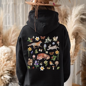 May include: Black hoodie featuring a colorful nature-themed design on the back. The design includes illustrations of foxes, rabbits, butterflies, flowers, and other woodland elements. The hoodie has a hood and long sleeves.
