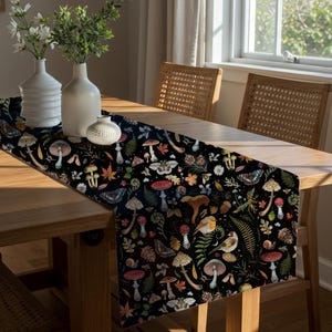 May include: A black table runner with a whimsical forest pattern featuring mushrooms, butterflies, and foliage. The runner is placed on a wooden table, with white vases and a small decorative item. The scene is illuminated by natural light.