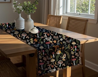 May include: A black table runner with a whimsical forest pattern featuring mushrooms, butterflies, and foliage. The runner is placed on a wooden table, with white vases and a small decorative item. The scene is illuminated by natural light.