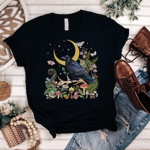 May include: A black t-shirt with a graphic print of a raven perched on a branch with a crescent moon and mushrooms. The raven is surrounded by flowers and leaves.