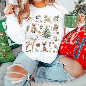May include: White sweatshirt with a festive winter scene featuring a variety of woodland animals, including a fox, deer, owl, and squirrel. The design also includes a Christmas tree, pinecones, and other holiday elements.