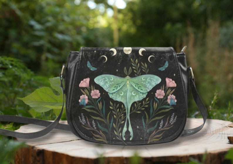 K&ouml;nnte beinhalten: Eine schwarze Leder-Umh&auml;ngetasche mit Mond- und Motten-Design. Die Tasche zeigt eine gr&uuml;ne Motte mit zarten Fl&uuml;geln, umgeben von Blumen und Bl&auml;ttern. Der Hintergrund ist dunkelblau mit einem Sternenhimmel-Effekt.