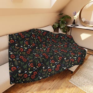 May include: A black throw blanket adorned with a floral design of red poppies, butterflies, and wildflowers. The blanket is draped over a white sofa, adding a decorative touch to the living room setting.
