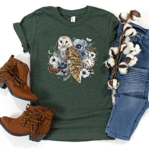 May include: A green t-shirt with a white barn owl surrounded by flowers and a pocket watch. The owl is perched on a branch with its wings spread. The flowers are in shades of pink, purple, and white.