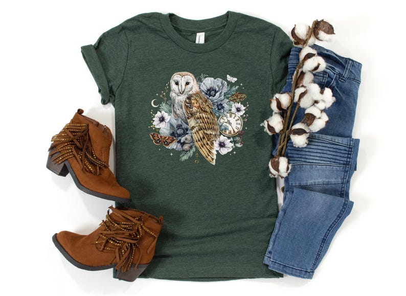 May include: A green t-shirt with a white barn owl surrounded by flowers and a pocket watch. The owl is perched on a branch with its wings spread. The flowers are in shades of pink, purple, and white.