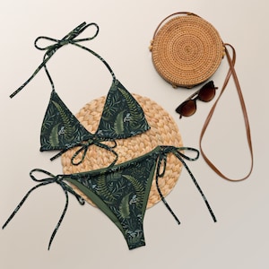May include: A green and black bikini with a leafy print. The bikini top is a triangle style with adjustable straps. The bikini bottom is a high-cut style with ties on the sides. The bikini is lying on a woven straw mat.