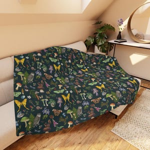 May include: A dark green throw blanket featuring a repeating pattern of mushrooms, butterflies, owls, and flowers. The blanket is draped over a white sofa, displaying the detailed, colourful design. A cosy home decor item.