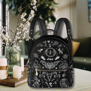 May include: Black backpack with a detailed white illustration of various occult symbols, including a moth, hands, and a skull. The backpack has adjustable straps and gold-colored zippers. The bag is sitting on a wooden surface.