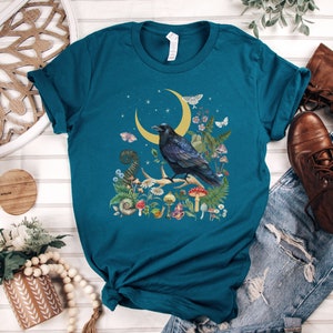 May include: Teal blue t-shirt with a black raven perched on a crescent moon surrounded by flowers and mushrooms.