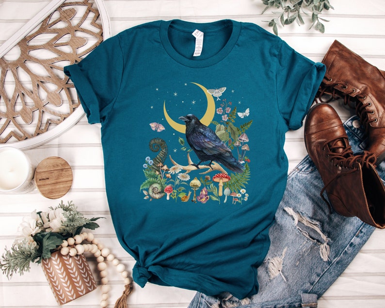 May include: Teal blue t-shirt with a black raven perched on a crescent moon surrounded by flowers and mushrooms.