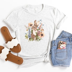Op de afbeelding: Een grijs T-shirt met een aquarel-illustratie van een bruin konijn omringd door kleurrijke bloemen. Het shirt wordt gecombineerd met een spijkerbroekje en witte sandalen.