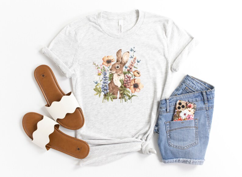 Op de afbeelding: Een grijs T-shirt met een aquarel-illustratie van een bruin konijn omringd door kleurrijke bloemen. Het shirt wordt gecombineerd met een spijkerbroekje en witte sandalen.