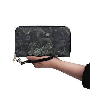 May include: A dark blue wallet with a dragon and floral pattern. The design features green dragons, white flowers, and crescent moons. The wallet has a black wrist strap and a gold zipper.