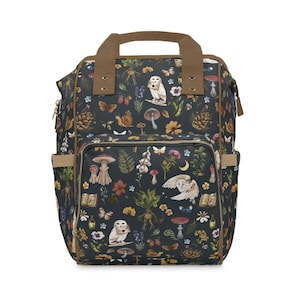 May include: A dark navy backpack with a whimsical forest pattern featuring owls, mushrooms, butterflies, and flowers. The backpack has brown accents on the handle and trim, with multiple pockets and a zippered closure.