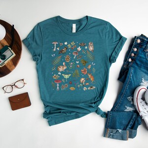 May include: A teal blue t-shirt with a white crew neck and a colorful illustration of woodland creatures, mushrooms, and plants. The shirt is laid flat on a white surface with a pair of jeans and white sneakers.