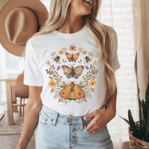 May include: A white short-sleeve t-shirt featuring a central graphic print of three butterflies arranged vertically, surrounded by a circular wreath of colorful wildflowers, small green leaves, and two bees on the upper left and right sides.
