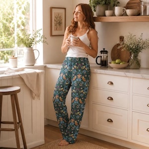 Pode incluir: Uma mulher usando calças de pijama verde-azulado com um padrão floral e de pássaros, combinadas com uma regata branca. Ela está segurando uma caneca em um ambiente de cozinha com um banquinho de madeira e uma janela ao fundo.