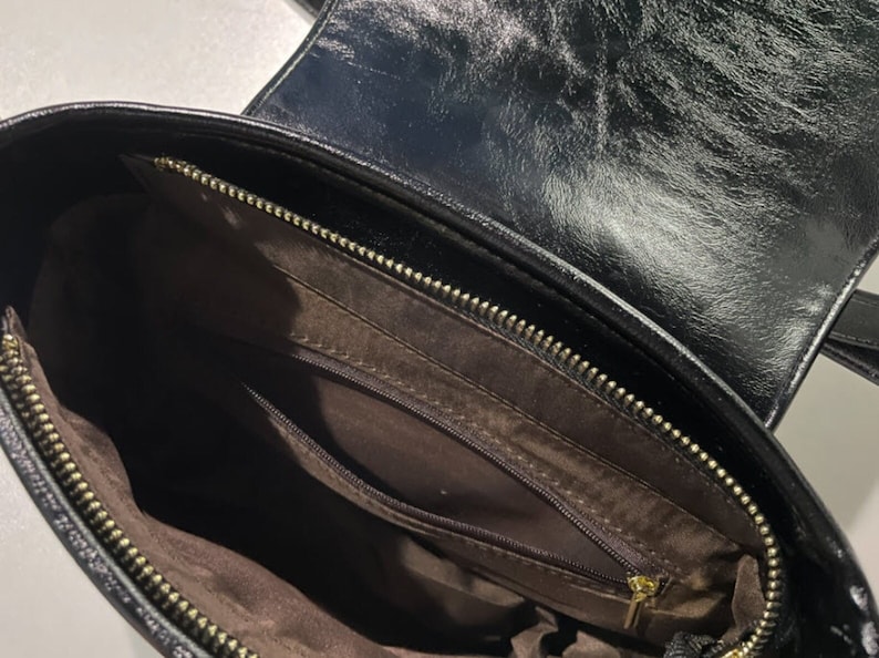 May include: Black leather handbag with a zippered pocket inside. The bag has a gold zipper and a brown lining.