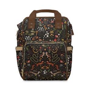 May include: A black backpack with a brown leather handle and straps. The backpack has a large pocket on the front with a zipper closure. The fabric of the backpack is covered in a pattern of mushrooms, flowers, owls, snakes, and other woodland creatures.