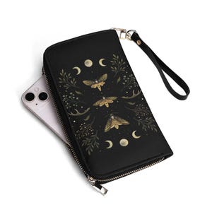 May include: Black wallet with a gold zipper, wrist strap, and a celestial design. The design features gold moths, crescent moons, and botanical elements. A smartphone is partially visible inside the wallet.