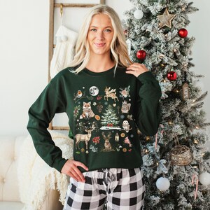 May include: A green Christmas sweater with a woodland scene featuring animals, plants, and a small Christmas tree. The sweater has a crew neck and long sleeves.