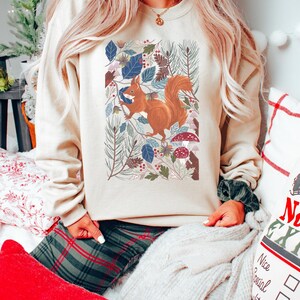 May include: A cream coloured sweatshirt featuring a graphic of a squirrel in a woodland scene. The squirrel is brown with a bushy tail. The woodland scene is filled with green leaves, pine needles, and red mushrooms.