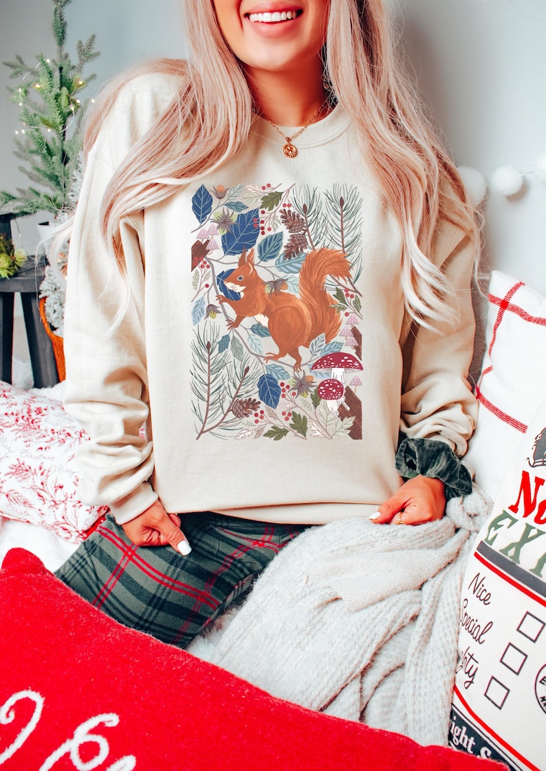 May include: A cream coloured sweatshirt featuring a graphic of a squirrel in a woodland scene. The squirrel is brown with a bushy tail. The woodland scene is filled with green leaves, pine needles, and red mushrooms.