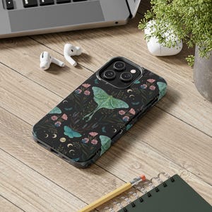 Puede incluir: Funda de teléfono negra con un diseño botánico que presenta una gran polilla verde, flores rosas y mariposas azules. La funda tiene un fondo oscuro con acentos de luna creciente y hojas. La funda del teléfono está sobre una superficie de madera.